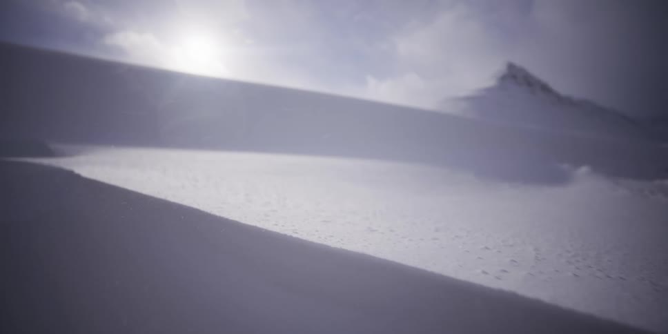 Wind sweeps across a sunlit snowdrift on a mountain in a dramatic slow-motion close-up