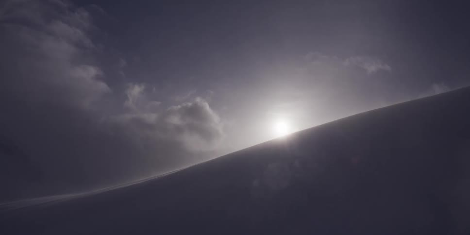 A bright sun shines over a dark, snowy mountain ridge as wind and snow blow across the desolate landscape