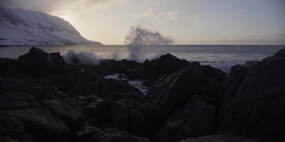 Slow motion footage of a powerful ocean wave crashing against a rocky shore with a snow-covered mountain in the background