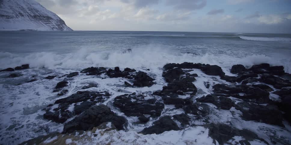 Slow-motion wide shot of powerful ocean waves crashing over snow-covered rocks on a winter coastline