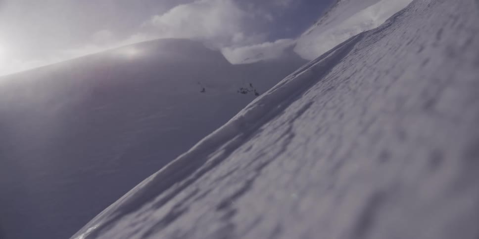 A low-angle close-up captures wind sweeping fine snow across a steep mountain face on a cold, bright day