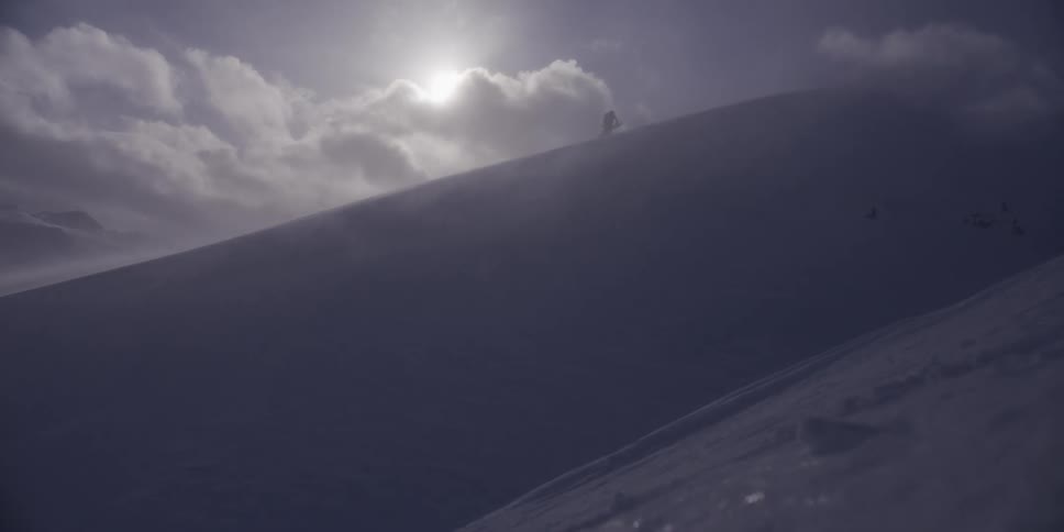 A solitary mountaineer slowly climbs a steep, snow-covered ridge during a dramatic blizzard as the sun shines through the clouds