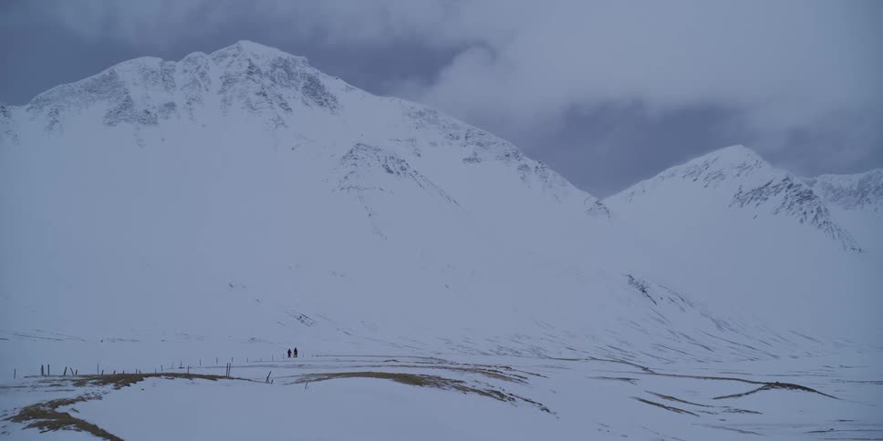 Two distant figures are dwarfed by the immense scale of a vast, snow-covered mountain landscape as they hike through the valley