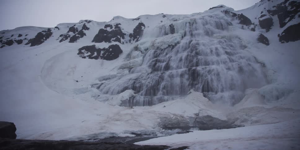 Water from a powerful melting glacier cascades down a snow-covered mountain range on a cold, overcast day