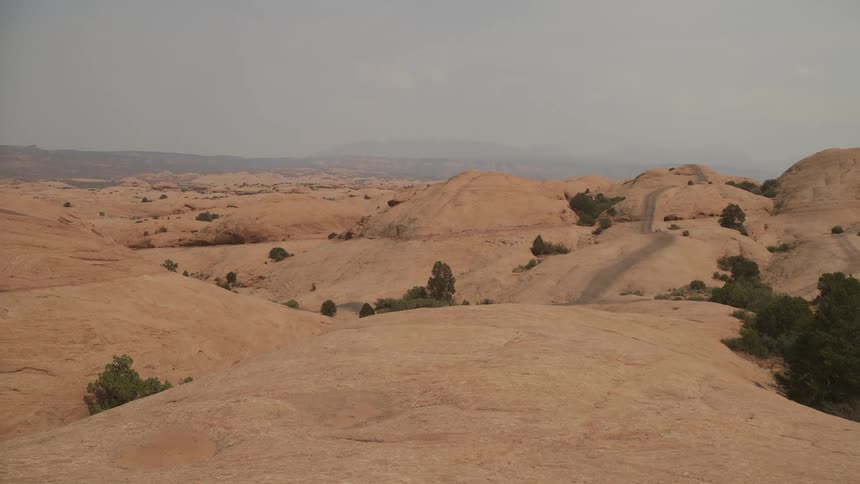 The rugged, rolling sandstone hills of a desert landscape stretch to the horizon under a hazy sky
