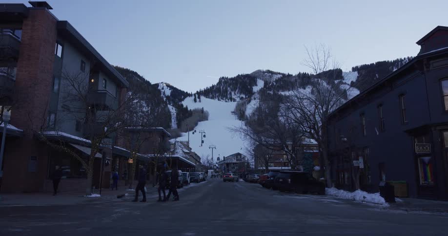 A wide shot of a classic ski resort town in Aspen, Colorado at dusk, with pedestrians crossing the street and the mountain slopes visible