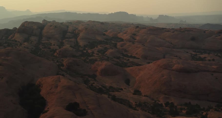 A vast, hazy landscape of red rock formations glows in the warm light of sunrise in a desert canyon
