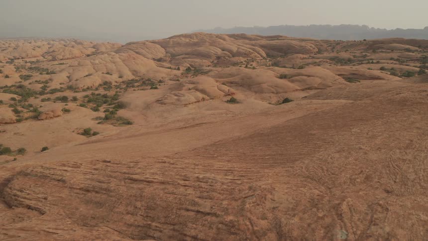 A vast and desolate landscape of rolling red slickrock formations stretches out under a hazy sky