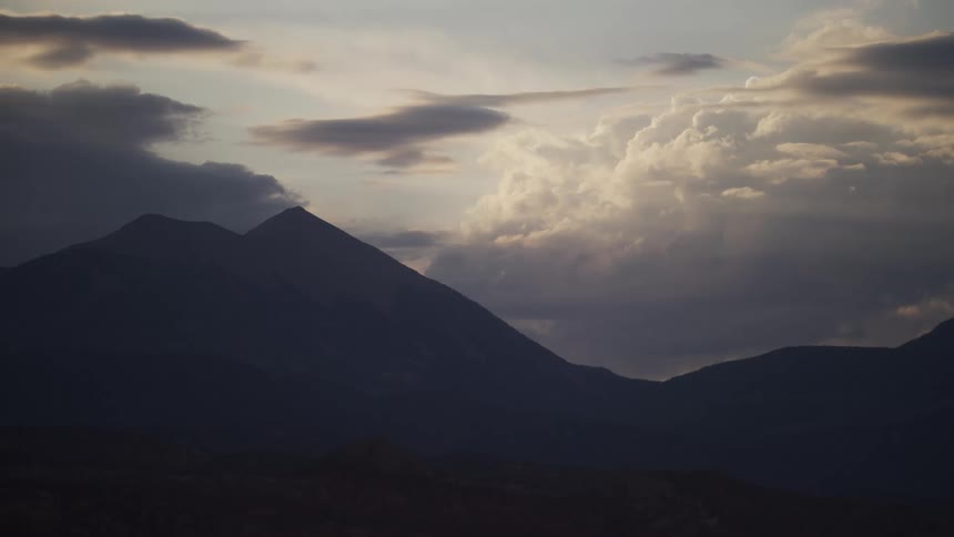 A silhouetted mountain range stands beneath a dramatic cloudy sky as sun rays peek through