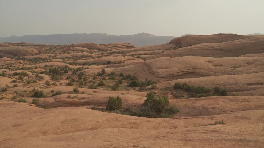 A wide, static shot captures the vast, arid expanse of rolling red rock formations and sparse desert vegetation under a hazy sky