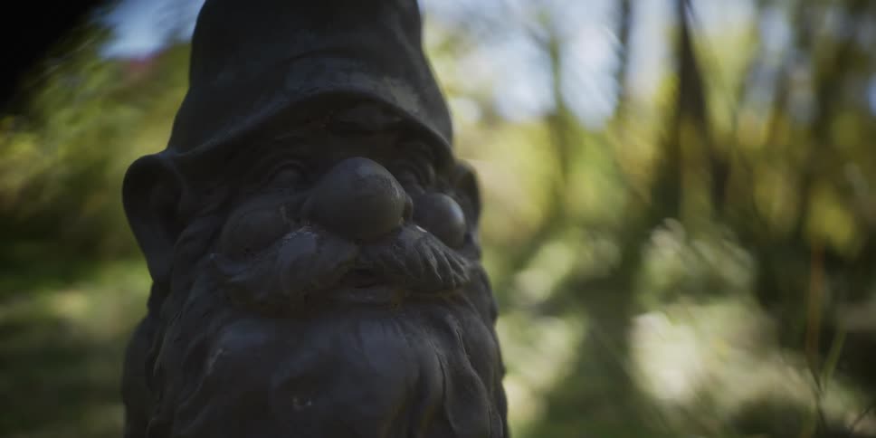 Static close-up shot of a weathered stone garden gnome with a blurry green background