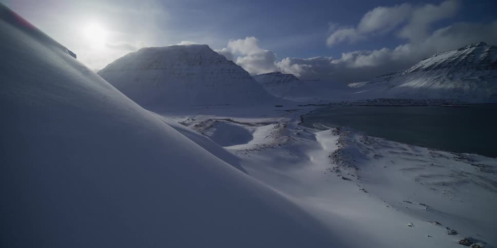 A vast, sunlit winter landscape shows snow-covered mountains surrounding a tranquil fjord