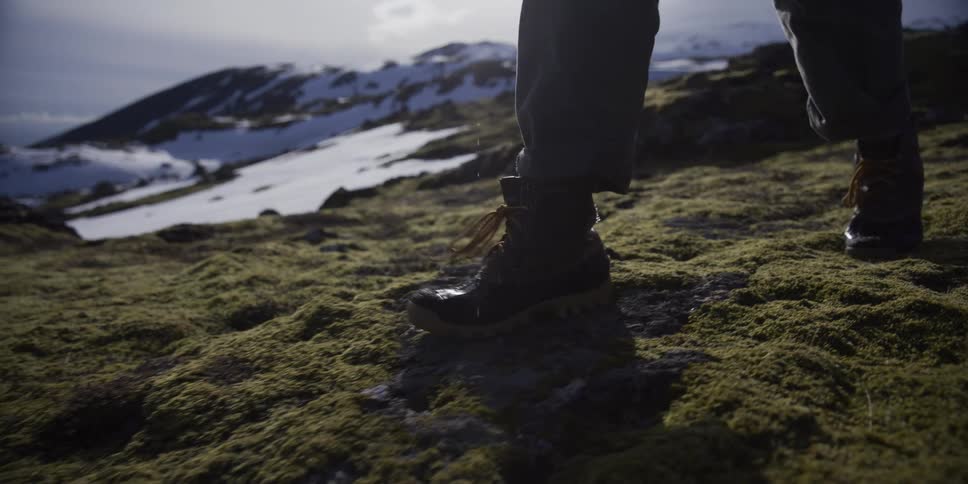 Close-up slow motion shot of a hiker's boots treading on lush green moss with sun flare and snowy mountains in the distance