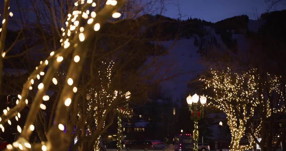 Festive holiday lights illuminate trees on a street in a cozy ski resort town at dusk with a snowy mountain in the background