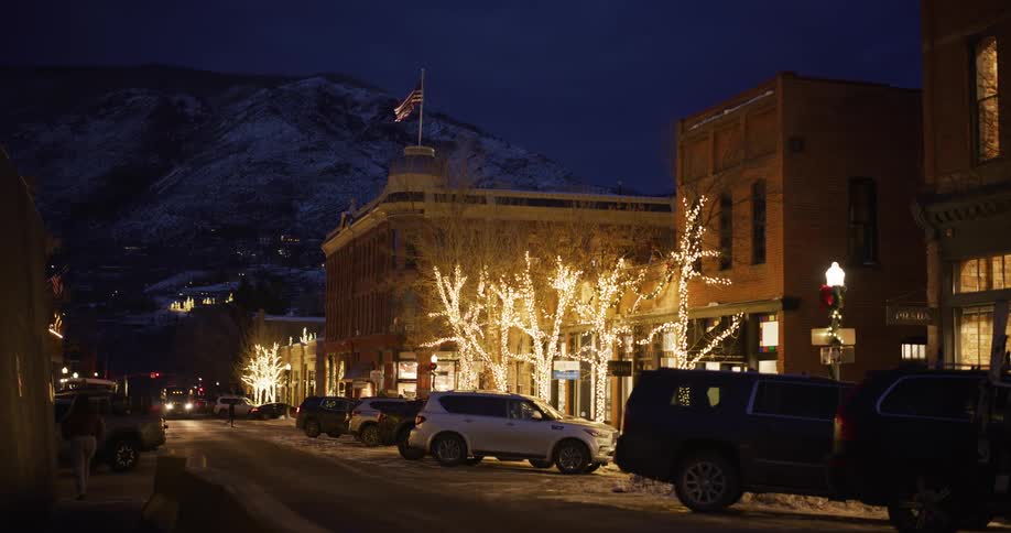 A quiet street in a snowy mountain town is decorated with festive Christmas lights at night