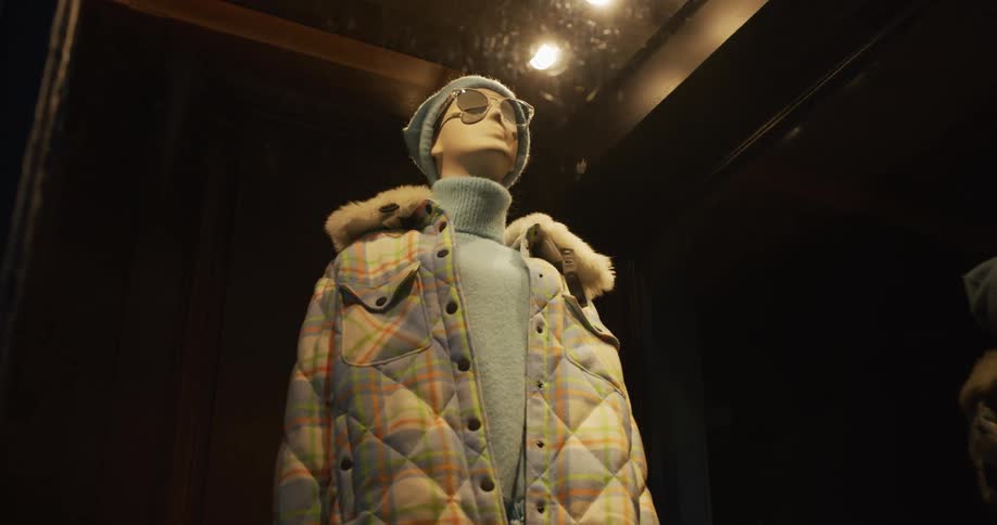 A low angle shot of a fashion mannequin wearing a winter coat, turtleneck, beanie, and sunglasses in a dark, spotlit store window display