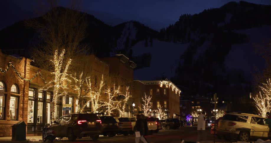 Festive Christmas lights illuminate a charming street in a snowy mountain town at night as people walk by