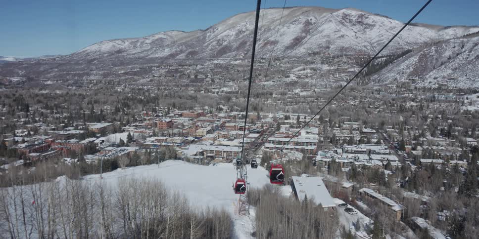 A scenic point-of-view from a ski gondola descending toward the snow-covered town of Aspen, Colorado, on a sunny winter day