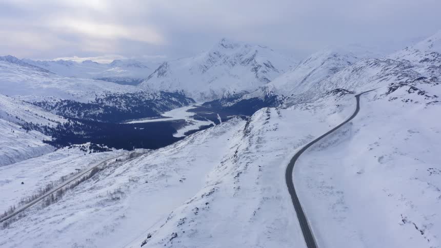 An empty highway winds through a vast, snowy Alaskan mountain range on a cold, overcast winter day