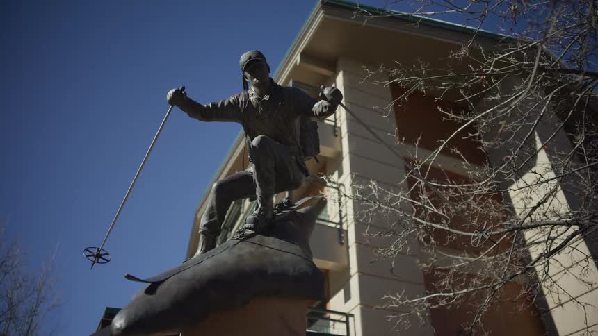 A low-angle shot of a bronze statue of a skier in front of a modern building with a clear blue sky