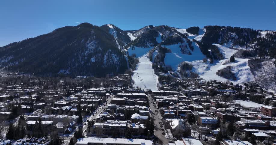 A wide, static aerial shot showcases the iconic ski town of Aspen, Colorado, with skiers descending the snow-covered Aspen Mountain on a bright, sunny day