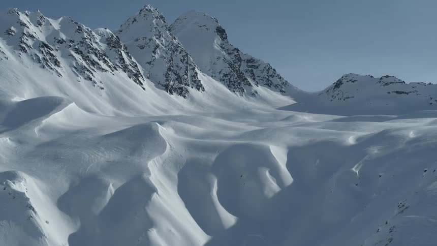 A snowboarder carves through fresh powder snow on a vast, sunlit mountain range