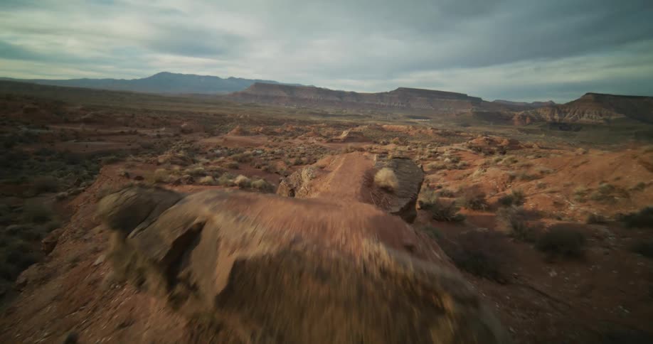 An FPV drone speeds across a vast red rock desert landscape, flying over unique rock formations and down into a sandy arroyo under a cloudy sky