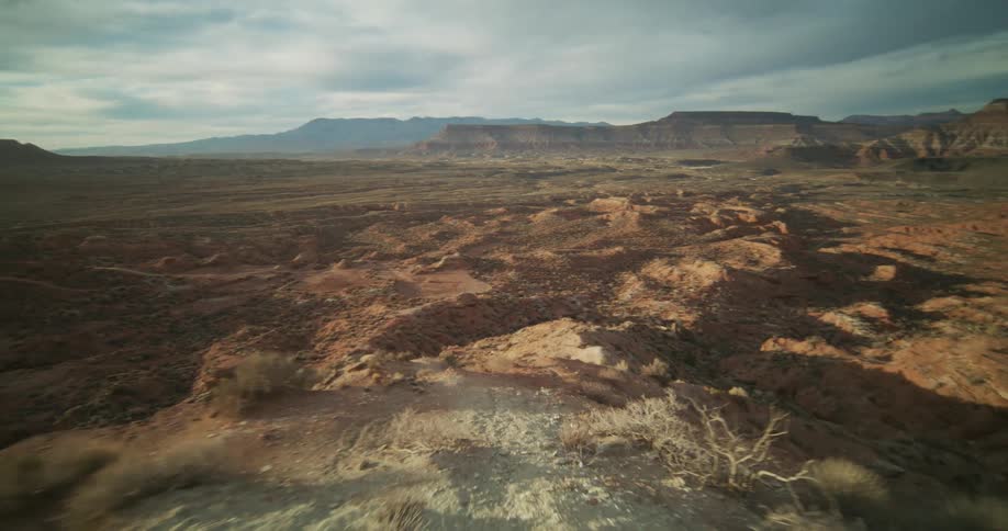 An aerial view swoops over the edge of a vast, arid red rock desert landscape under a cloudy sky