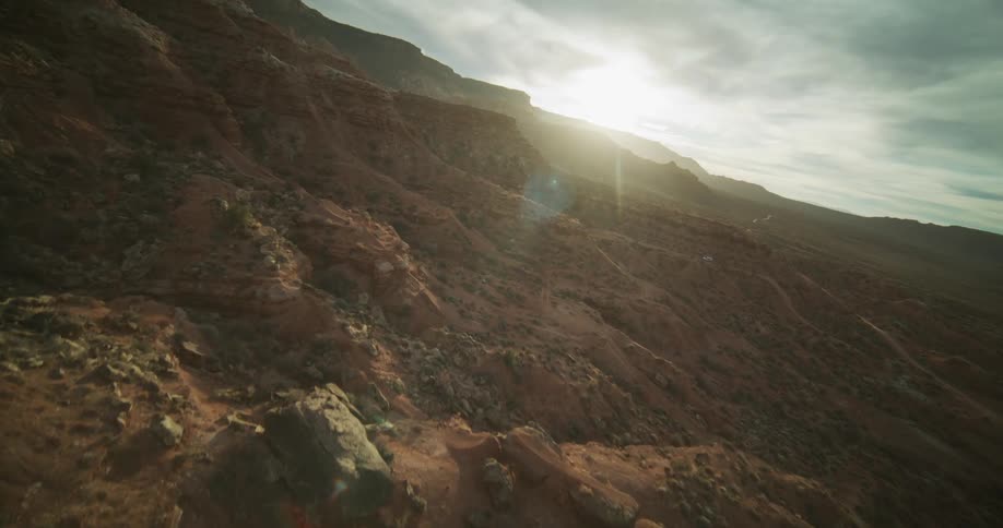 An aerial drone slowly flies up the side of a vast red rock canyon as the sun sets in the distance