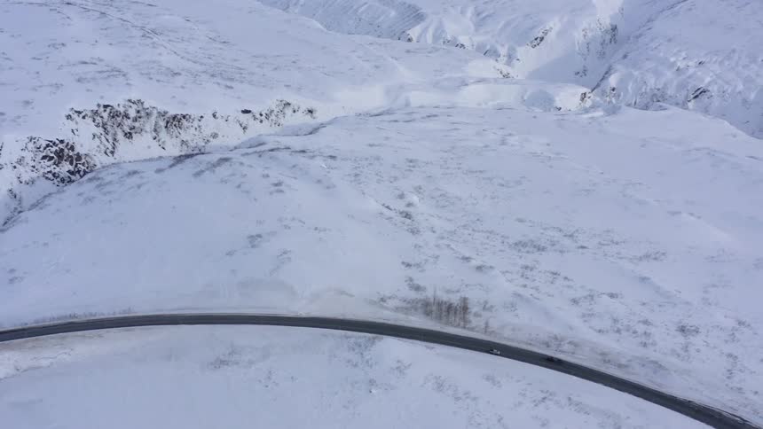Aerial view of two cars driving along a winding mountain road through a vast, snow-covered winter landscape