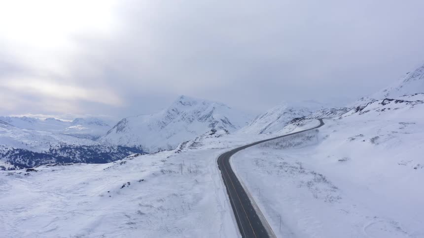 An aerial view shows a lone car driving down a winding road through a vast, snow-covered mountain landscape in winter
