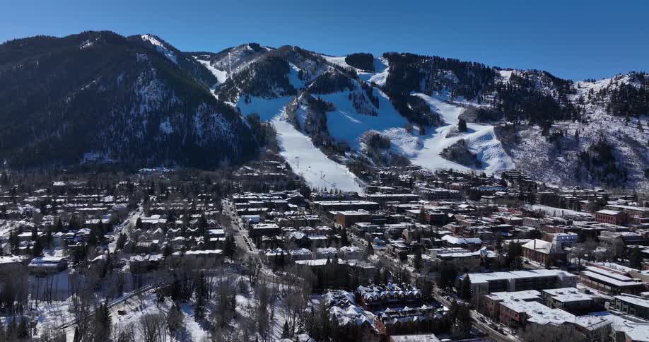 An aerial view pulls back to reveal the beautiful ski resort town of Aspen, Colorado, at the base of a vast, snow-covered mountain on a sunny day