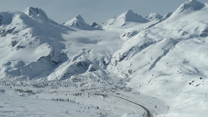 An expansive aerial view of the Worthington Glacier and a winding highway in a vast, snow-covered valley surrounded by mountains in Alaska