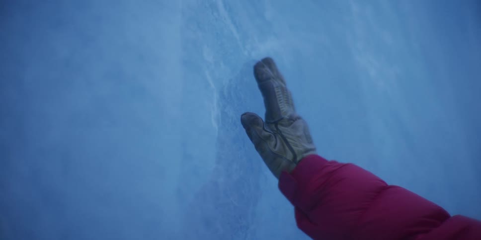 From a first-person perspective, a gloved hand gently touches and slides across a vast wall of blue glacial ice