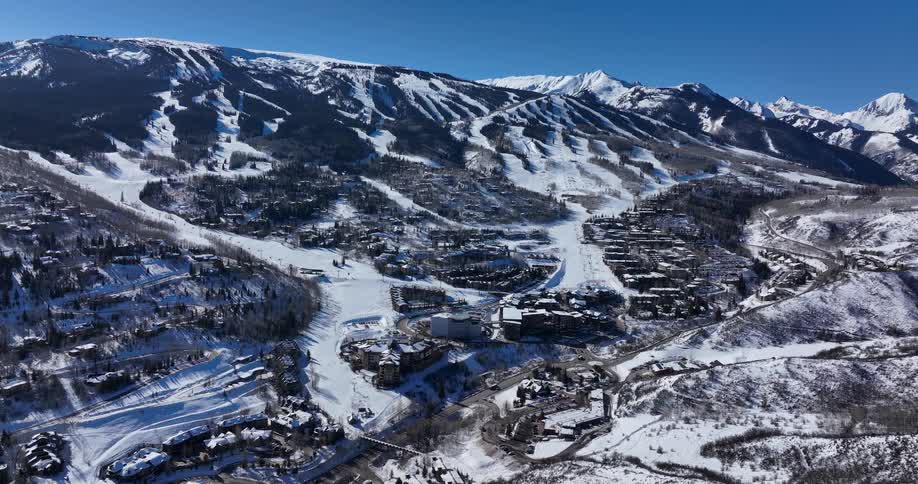 A wide aerial view shows a sprawling ski resort village nestled at the base of a snow-covered mountain on a bright, sunny day