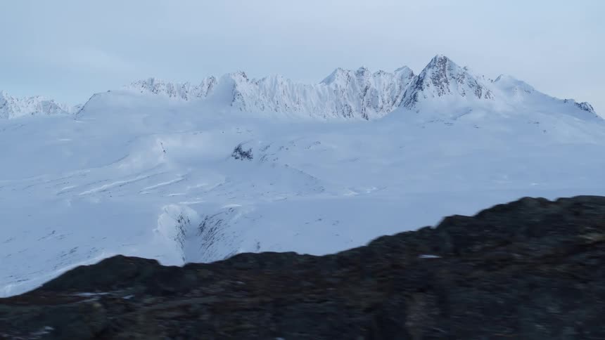 Slowly tracking across a frozen, rocky foreground to reveal a majestic, snow-covered mountain range in a vast and desolate landscape