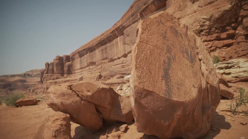 A slow focus pull reveals a large red rock boulder in front of a majestic desert canyon wall on a sunny day
