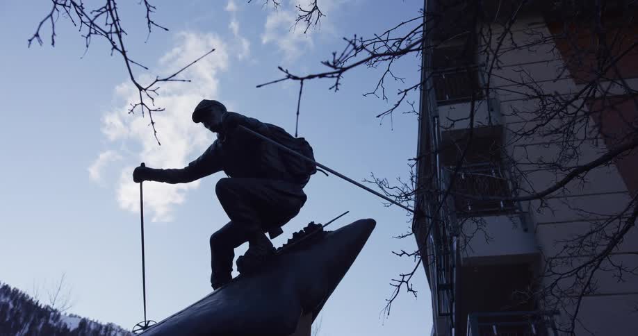 A slow zoom out reveals the silhouette of a bronze skier statue against a blue sky, framed by bare winter branches