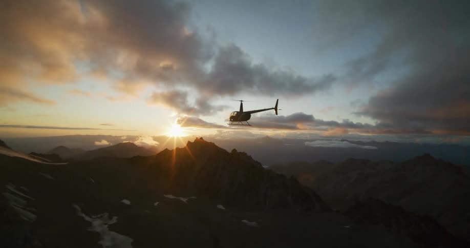 A silhouetted helicopter flies over a dramatic mountain range as the sun sets on the horizon