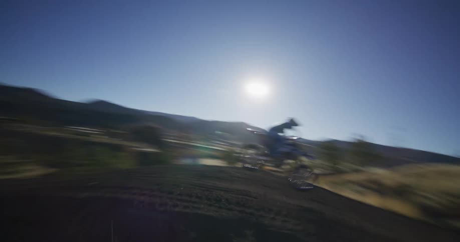 A dynamic FPV drone shot captures a motocross rider jumping a large dirt mound in slow motion on a sunny day