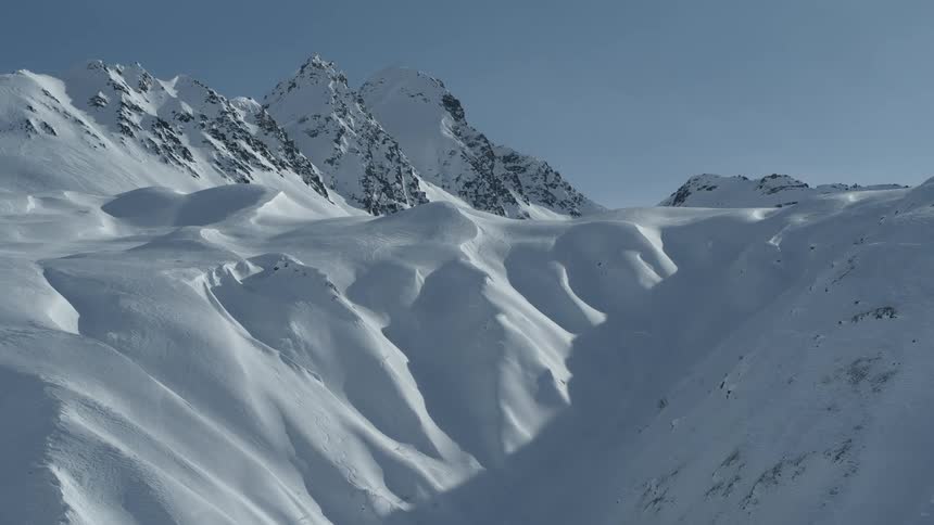 A slow aerial pullback reveals a vast and pristine snow-covered mountain range under a clear blue sky