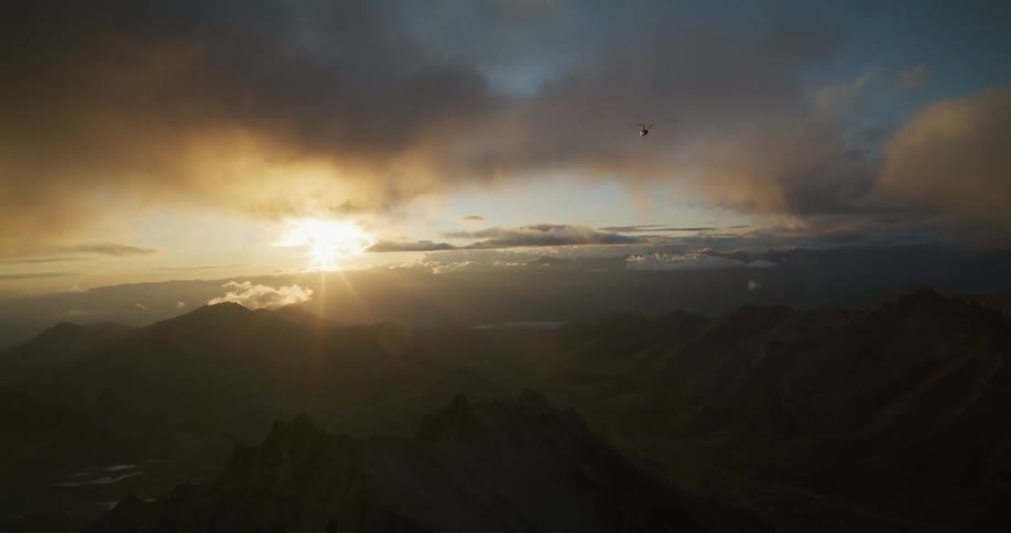 A helicopter flies towards the camera over a majestic mountain range during a golden sunset