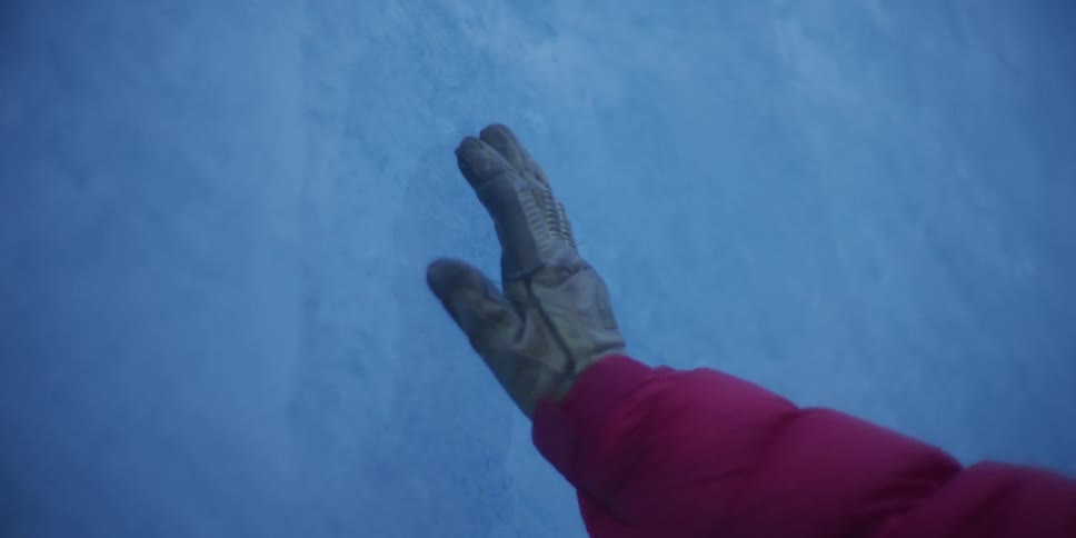 A first-person perspective shot of a gloved hand gently sliding across the textured blue ice wall of a glacier