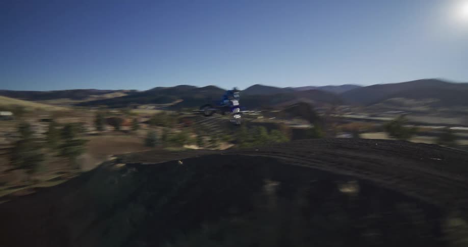 A motocross rider in full gear launches off a large dirt jump in slow motion on a sunny day, with rolling hills in the background
