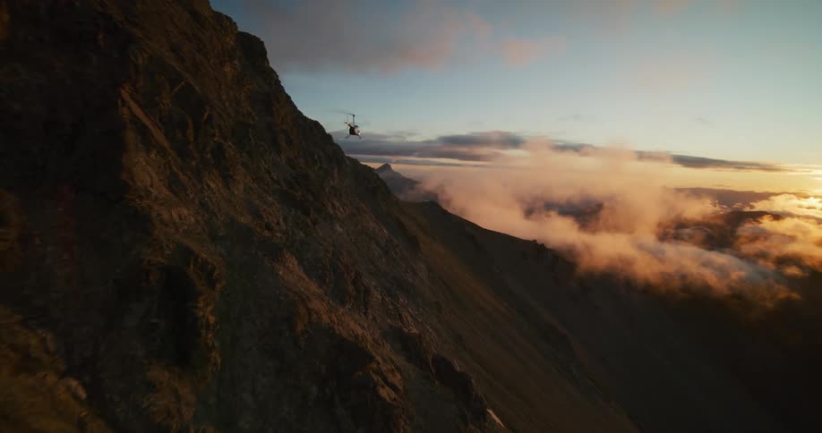 A helicopter takes off from a rugged mountain ridge, flying into a breathtaking sunset above the clouds