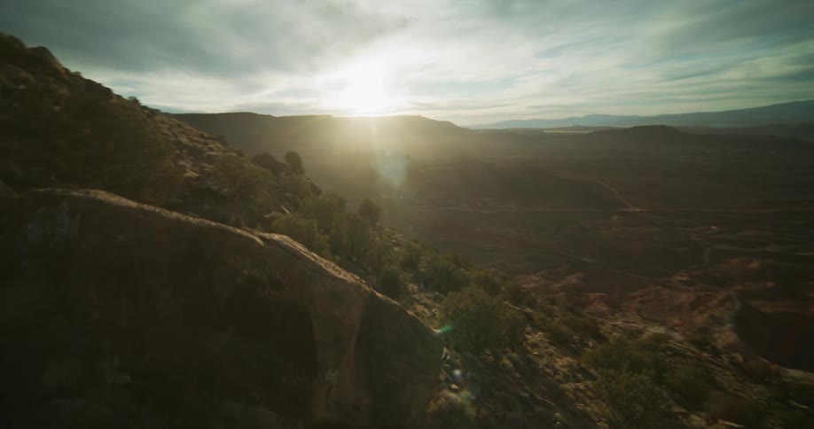 An epic aerial shot flies over a rocky mountainside to reveal a vast desert landscape glowing in the morning sun