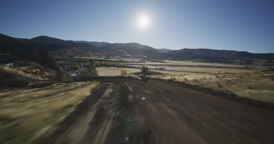 FPV aerial follows a motocross rider speeding along a dirt track in a sunny mountain valley