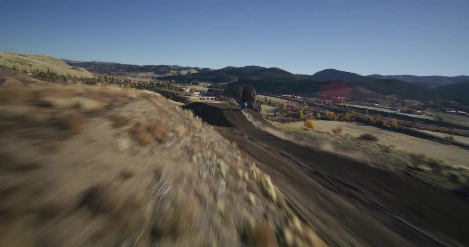 Dynamic aerial shot follows a motocross rider racing down a steep dirt track in a vast, sunny landscape