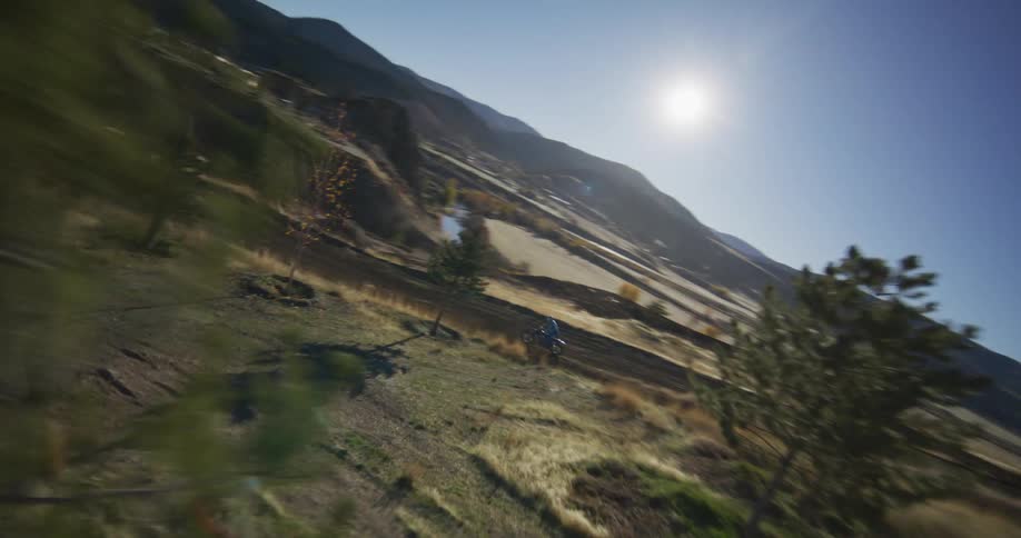 An aerial view follows a motocross rider speeding down a dusty track on a sunny day in the mountains
