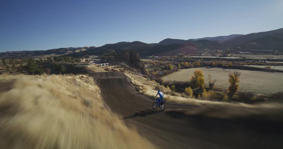 A high-speed FPV drone follows a motocross rider performing impressive jumps and turns on a professional dirt track in a beautiful mountain landscape